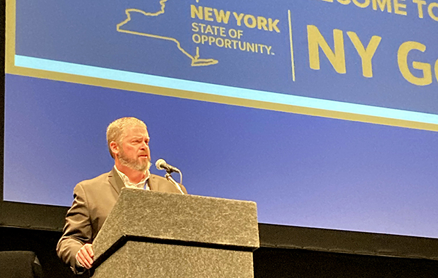A white man stands at a podium. Behind him is the New York state logo