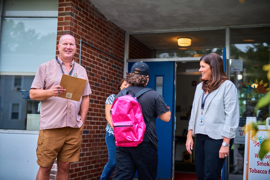 Two adults smile as students enter a school building