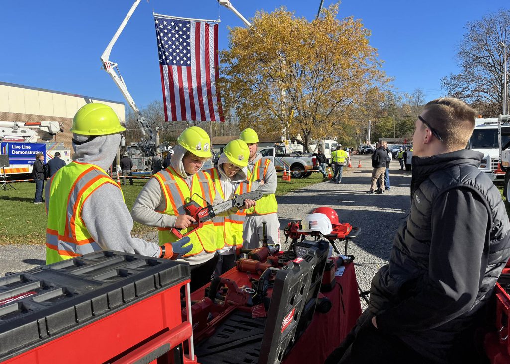 Students wearing safety gear show off a chainshaw. Behind them, a large American flag hangs from a utility truck. They are outdoors. 