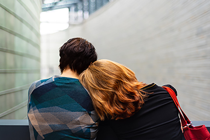 Two people are seen from behind. One is leaning their head on the other's shoulder