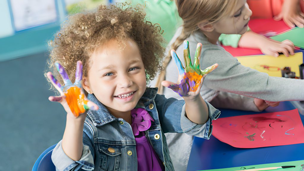 A young student with curly hair holds up hands covered in paint while smiling