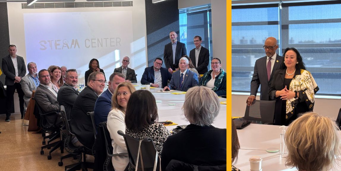 Several people sitting around a long rectangular table with a presentation screen on the front wall and 2 people standing up facing everyone sitting at the long table.