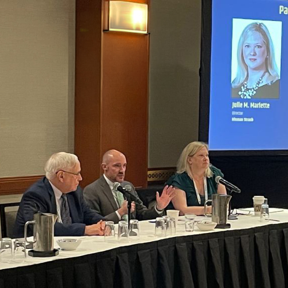Three people sitting at a table with microphones to speak. A face and title are on a presentation screen off to the side and behind them.