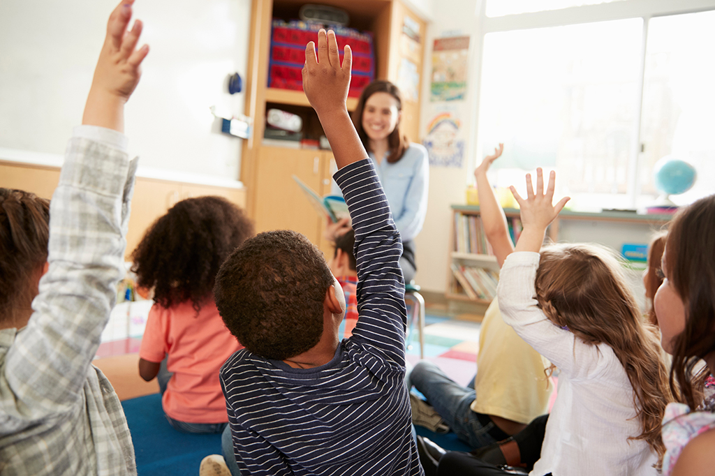 Students raise their hands while a teacher smiles.