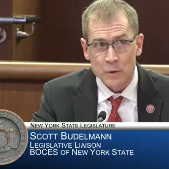 A person is facing forward sitting behind a table to speak.