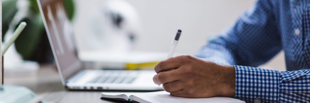 closeup of a man's hand writing with a laptop in the background.