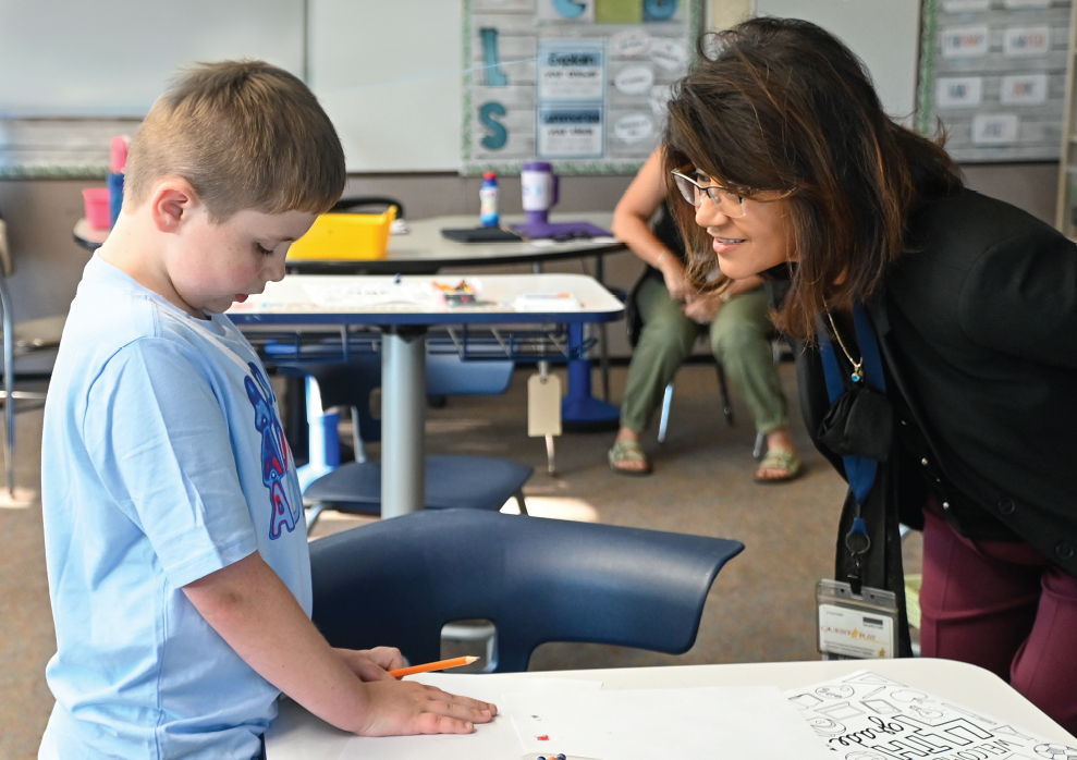 A teacher leans down to talk to a student in a classroom setting.