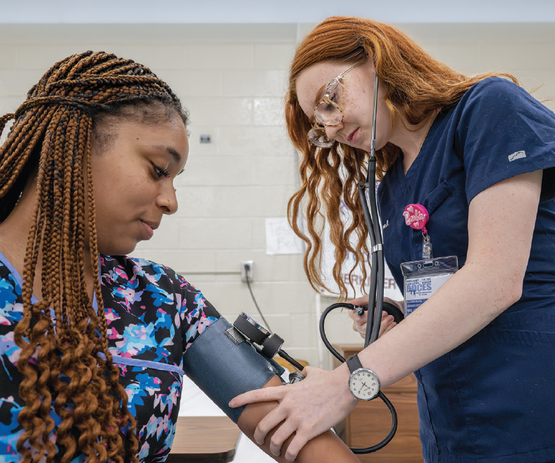A student in scrubs takes the blood pressure of another student in scrubs.