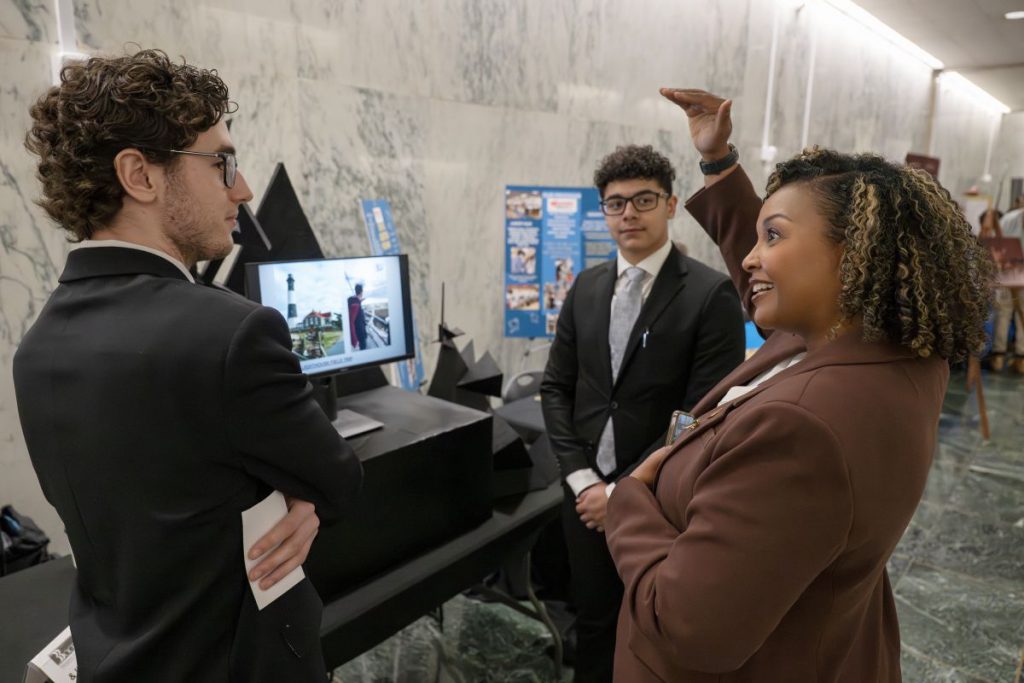 Two young men in dark suits stand talking with a woman in a brown suit.