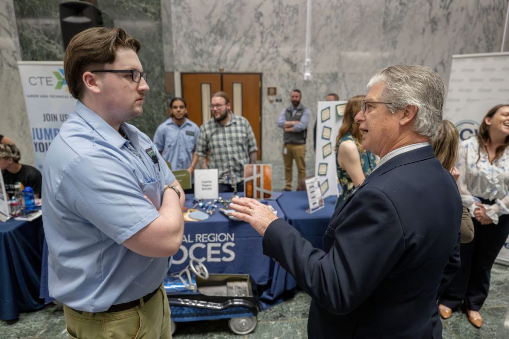 A male auto body student in a light blue polo stands arms crossed speaking with an older man in a navy suit
