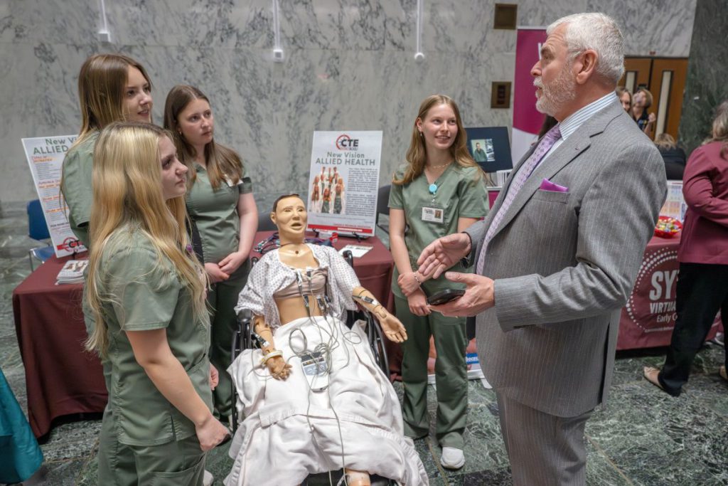 Four female Allied Health students in scrubs talk with a male legislator around a dummy with various health monitors attached to it
