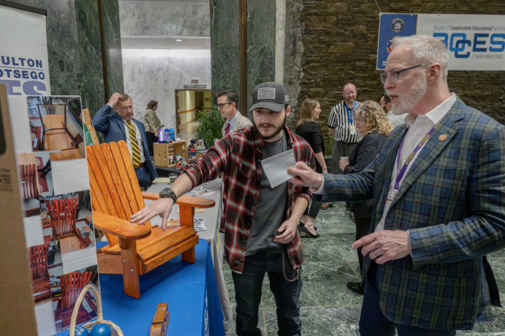 A woodworking student points to a miniature Adirondack chair while talking to a district superintendent in a plaid suit