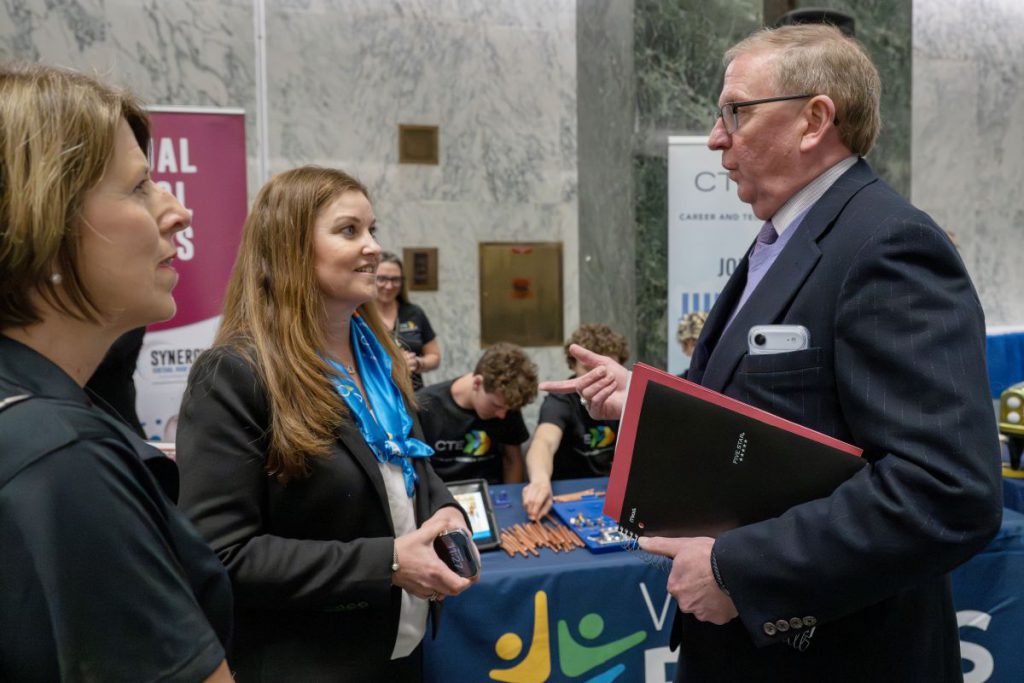 Two females with brown hair talk with an older male with glasses dressed in a navy suit
