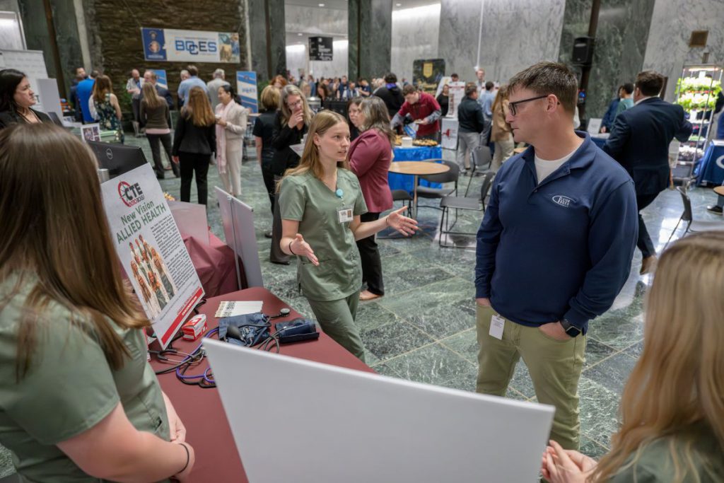A blond student in green scrubs stands with arms outstretched talking to a male in a blue shirt and khakis