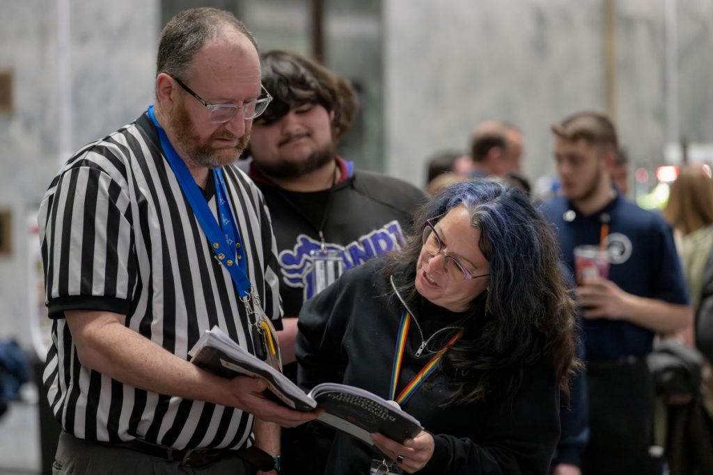 An bearded man in a black and white striped referee shirt stands next to a shorter woman with blue streaked dark hair and a rainbow lanyard, holding an open program book